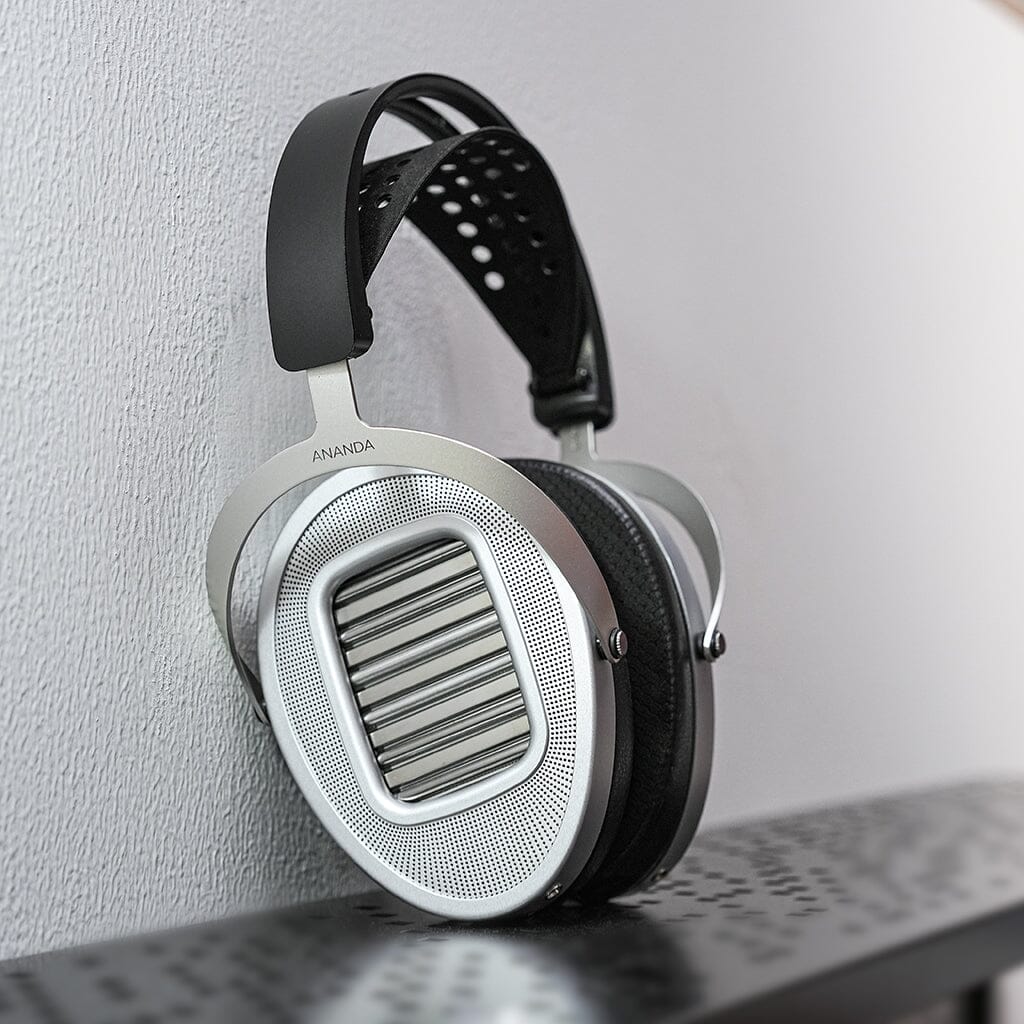 A pair of Hifiman Ananda Unveiled headphones with silver ear cups and a black headband hangs on the edge of a perforated metal surface near a textured light gray wall.