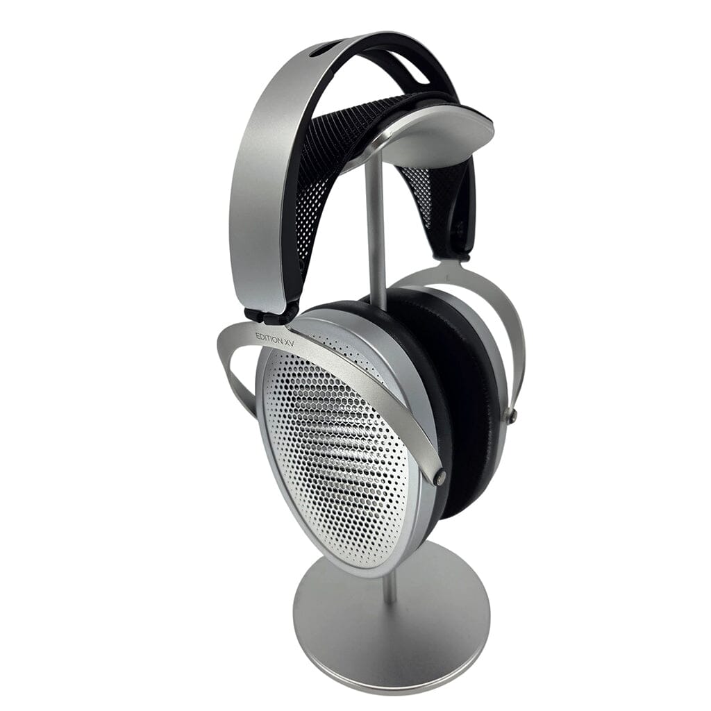 A pair of silver and black Hifiman over-ear headphones with mesh ear cups, featuring the Capra Audio Capra Comfort Strap for support, rests on a sleek metallic stand against a white background.