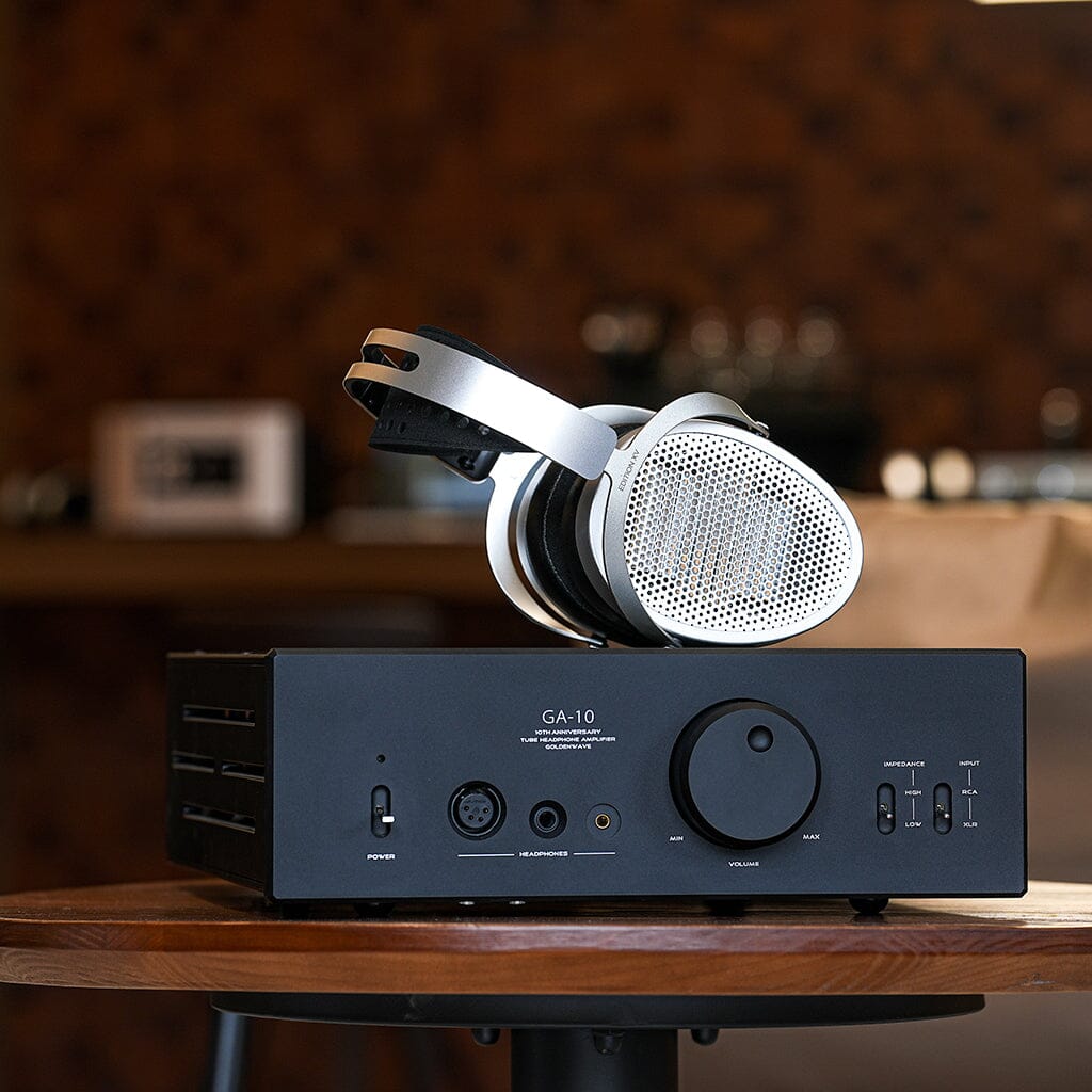 A pair of Hifiman Edition XV Headphones with NEO Supernano Diaphragm in silver and black rests on a black audio amplifier with multiple knobs and ports, all set on a wooden table against a blurred background.