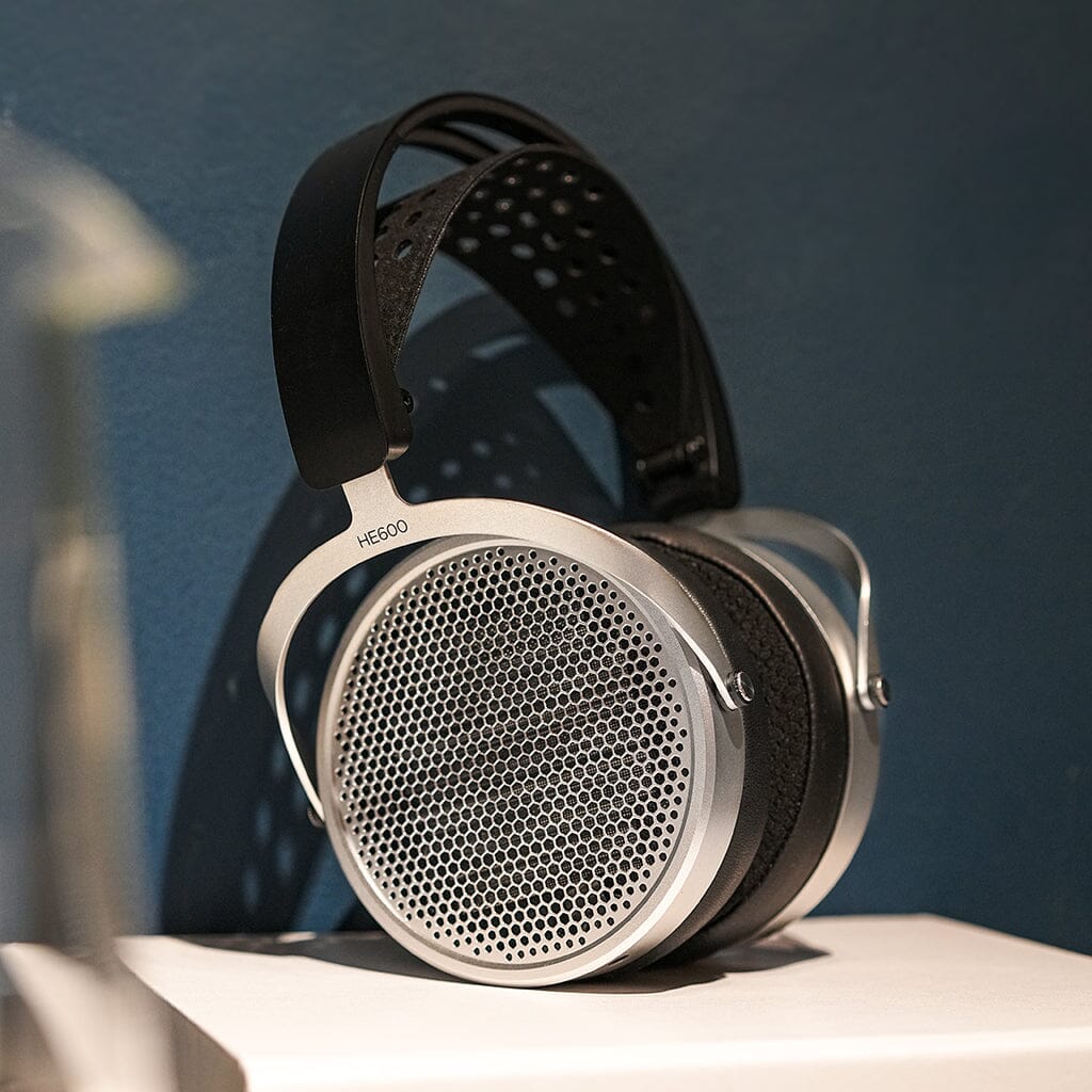 Close-up of Hifiman HE600 over-ear planar magnetic headphones with a perforated metal grill and black padded headband, displayed on a white surface with a blue background. Brand: Hifiman.