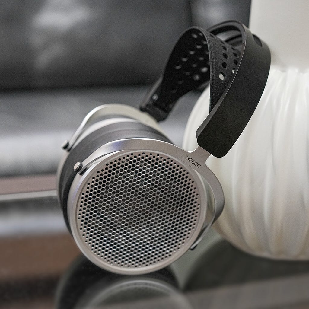 The Hifiman HE600 Headphones by Hifiman, featuring perforated ear cups, rest against a white vase on a reflective surface with a blurred couch in the background—ideal for use with high-quality headphone amplifiers.