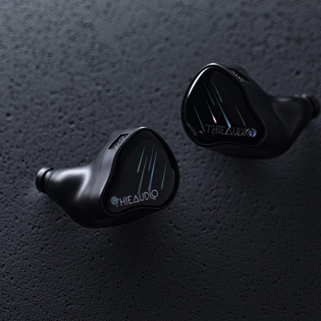 The Thieaudio Monarch MKIV In-Ear Headphones by Thieaudio rest on a textured dark surface, featuring a sleek, curved black design with subtle reflective lines and an adjustable tuning switch on the faceplates.