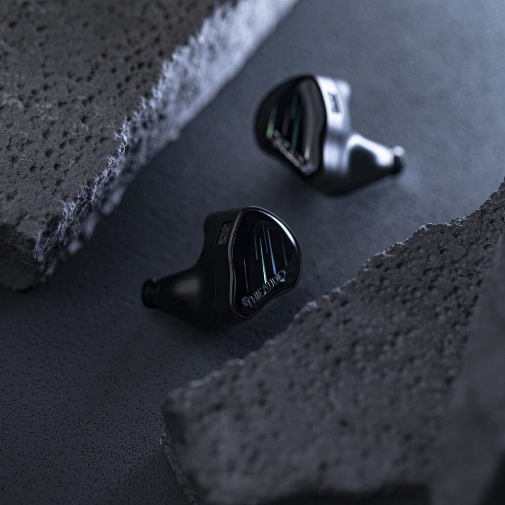 Two black Thieaudio Monarch MKIV In-Ear Headphones rest on a textured dark surface between rough gray concrete, illuminated by soft light that highlights their sleek, reflective design and adjustable tuning switch.