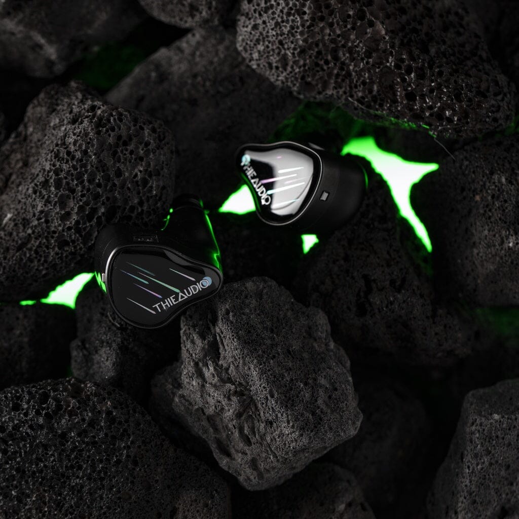 A pair of Thieaudio Monarch MKIV In-Ear Headphones sits on rugged black volcanic rocks, illuminated by a glowing green light that accentuates their precise design and adjustable tuning switch.