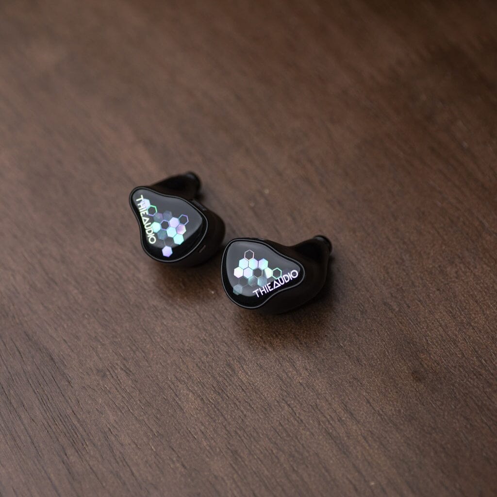 A pair of Thieaudio Monarch MKIV In-Ear Headphones with a black geometric hexagon design and an adjustable tuning switch for personalized sound rest on a dark wooden surface.