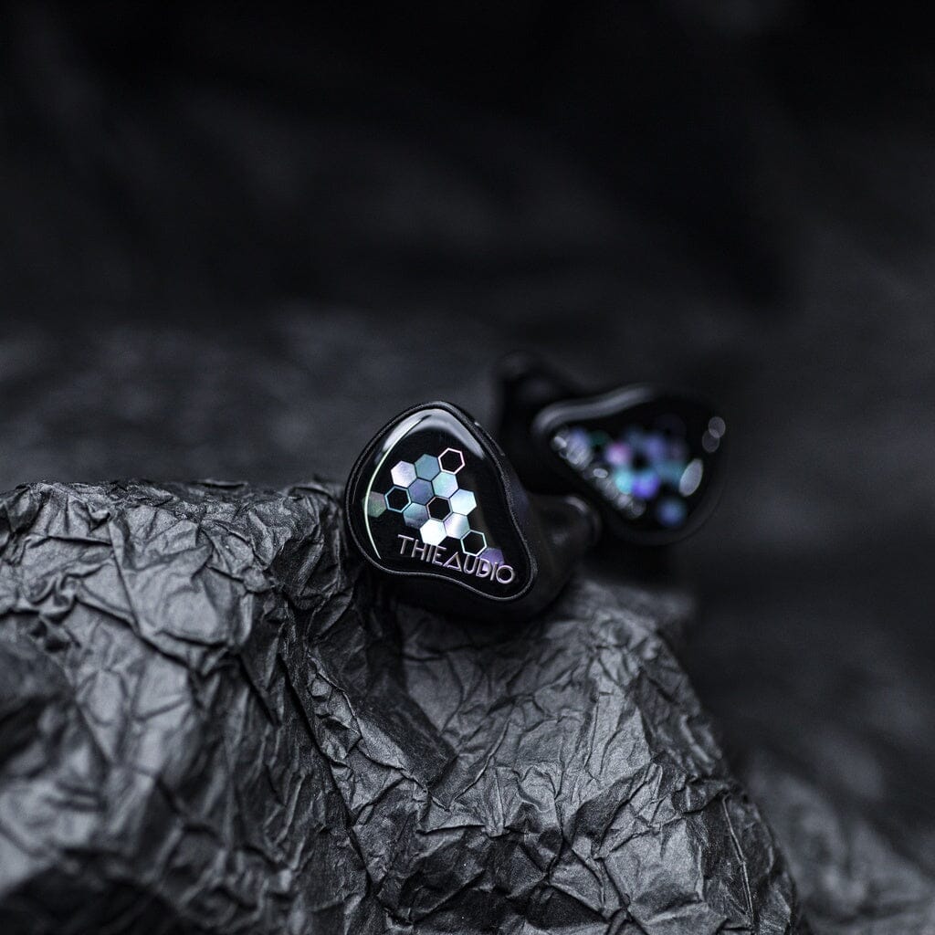 A pair of Thieaudio Monarch MKIV In-Ear Headphones with a black hexagonal pattern and an adjustable tuning switch, resting on crumpled black paper.