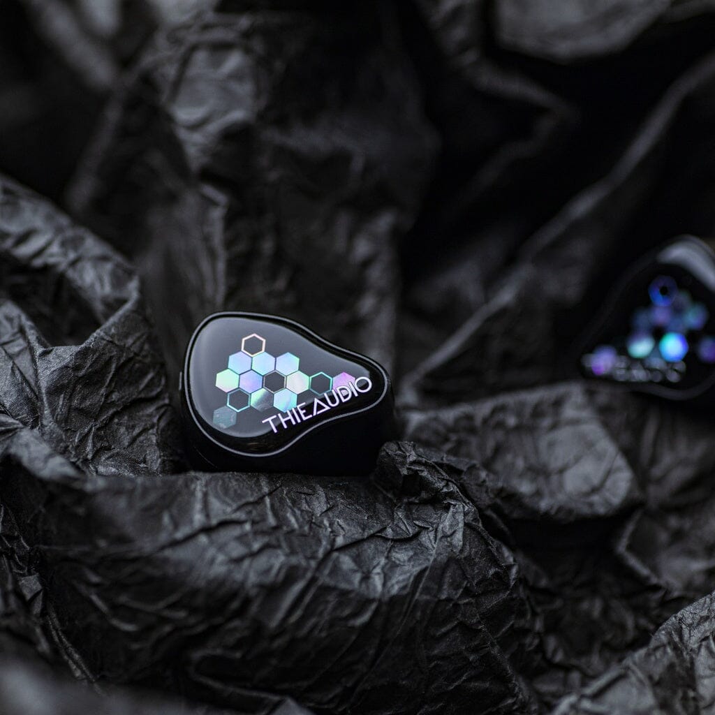 A close-up of Thieaudio Monarch MKIV In-Ear Headphones features a geometric hexagon pattern and logo, placed on textured black crumpled material.