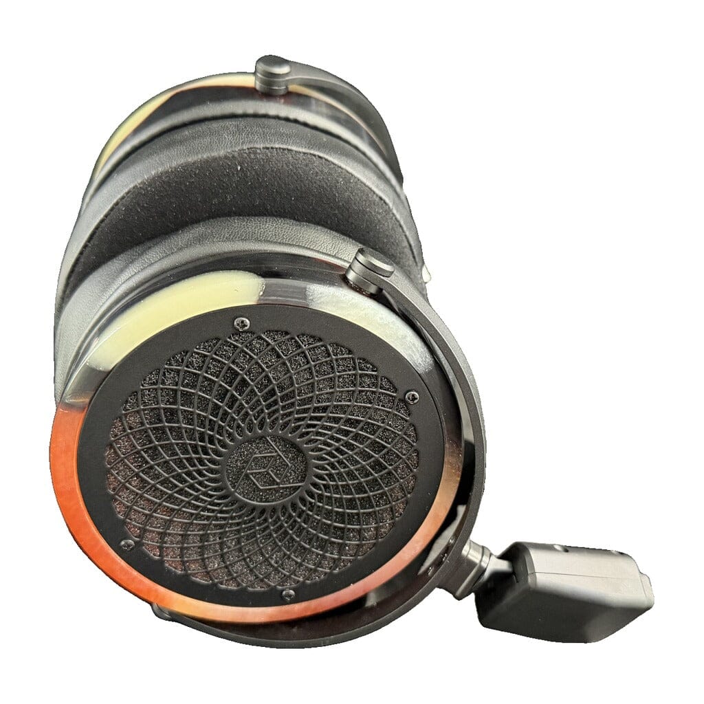 A close-up of a large, round, black earpiece from the Rosson Audio Design RAD-0 Headphones (Open Box C Condition - Clearance - 74), featuring a striking geometric grille pattern, attached cable, and visible metallic and orange accents along the sides, isolated on a white background.