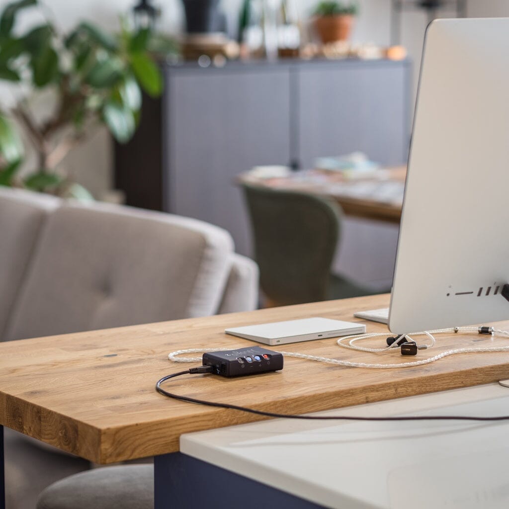 A modern desk with a computer monitor, keyboard, and the Chord Electronics Mojo 2 Portable DAC and Headphone Amplifier (4.4mm) sits in a bright room with plants, chairs, and a blurred cabinet in the background.