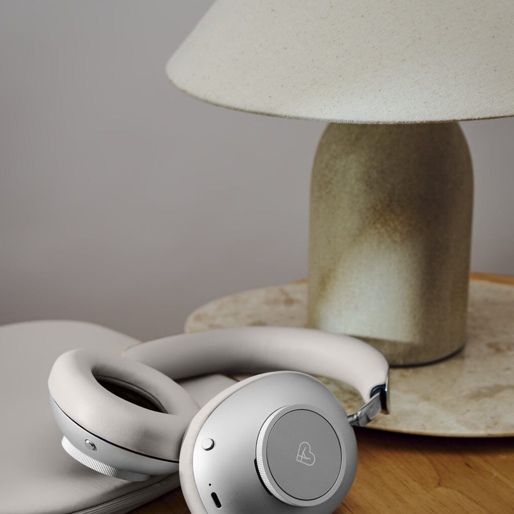 A pair of Loewe Leo Wireless Headphones with adaptive noise cancellation rests on a closed laptop next to a beige table lamp on a wooden surface.