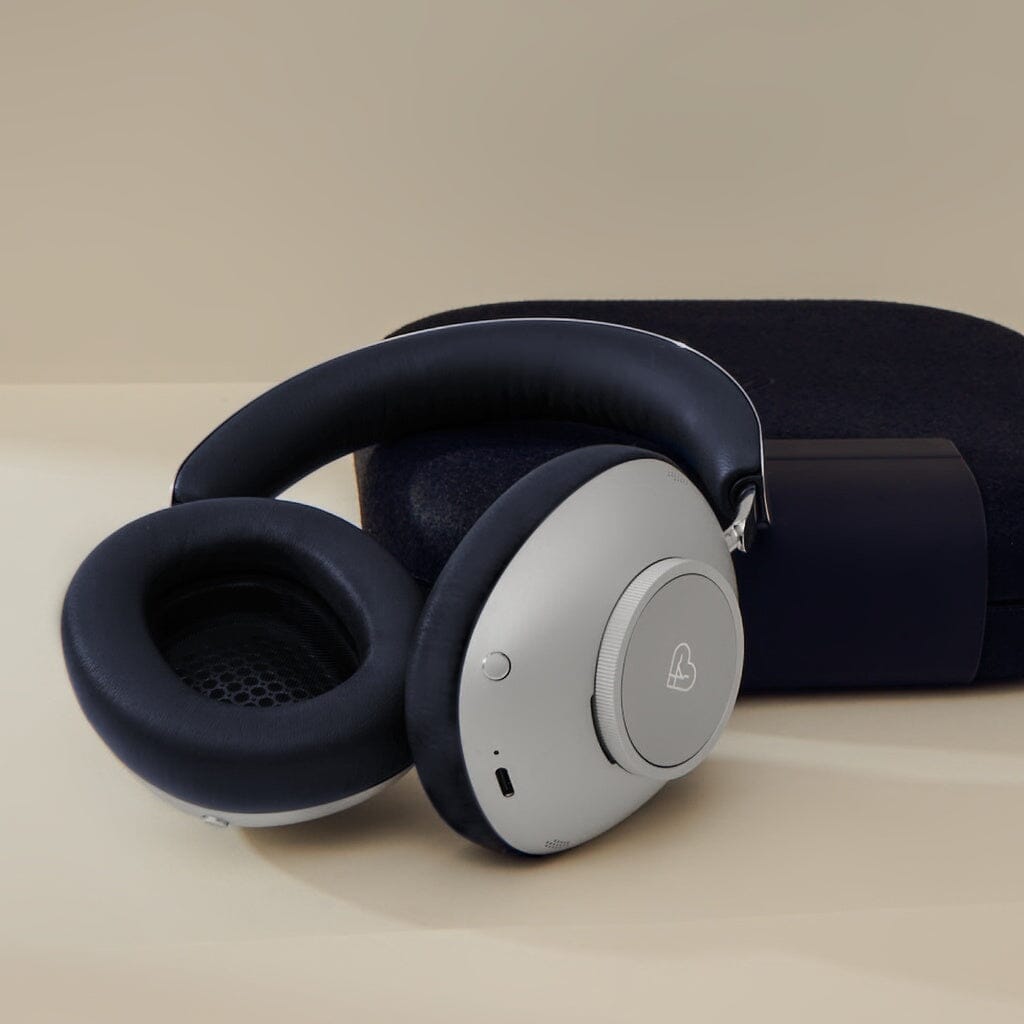 The Loewe Leo Wireless Headphones by Loewe feature a sleek, premium over-ear design in silver and black with adaptive noise cancellation, shown resting next to a matching dark carrying case on a beige surface.