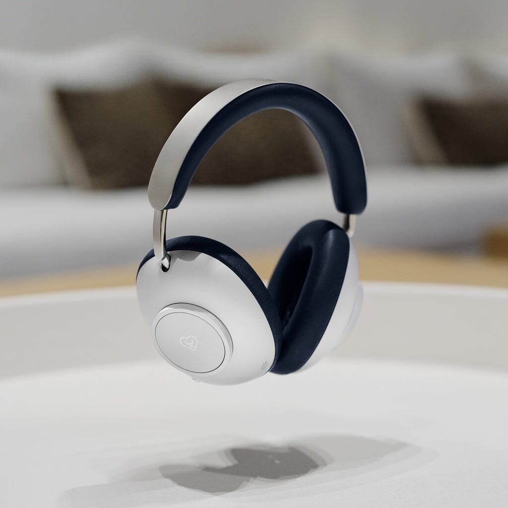 The Loewe Leo Wireless Headphones by Loewe, featuring adaptive noise cancellation, hover above a white surface with their modern over-ear design, white ear cups, and dark blue headband visible; a blurred couch and pillows appear in the background.