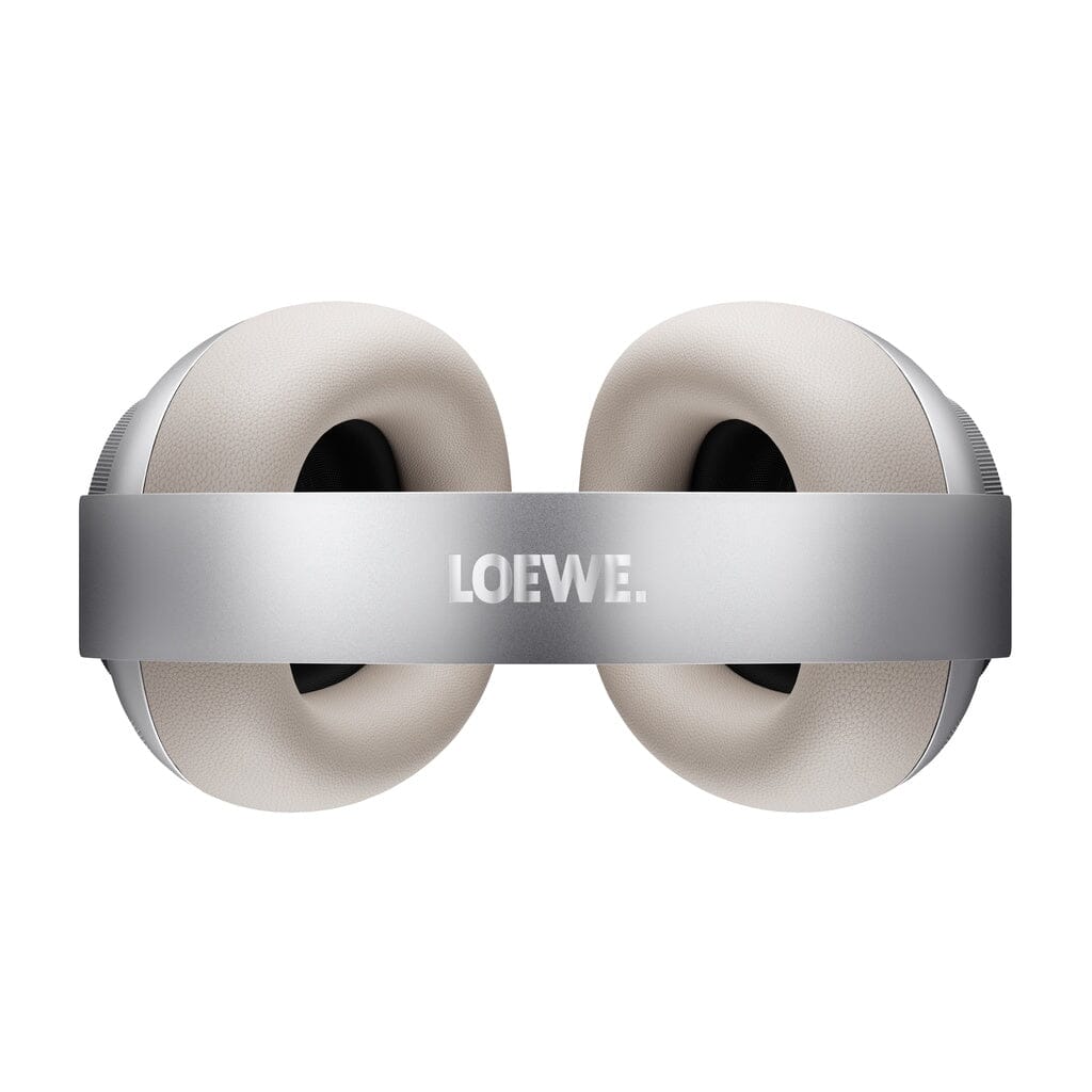 The Loewe Leo Wireless Headphones by Loewe are shown from a top-down perspective on a white background, featuring a silver design with large, cushioned beige ear pads and a metallic headband adorned with the LOEWE logo. These headphones provide adaptive noise cancellation for an immersive and personalized sound experience.