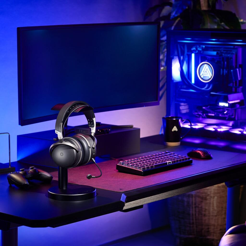 A striking gaming setup features a monitor, mechanical keyboard, mouse, and the Audeze Maxwell 2 ANC Wireless Headphones by Audeze displayed on a stand—all highlighted alongside a desktop PC aglow with purple lighting, set atop a dark desk in a blue-lit room. This immersive environment is ideal for hours of uninterrupted gameplay, elevated by the active noise cancellation of the Audeze Maxwell 2 that keeps distractions at bay.