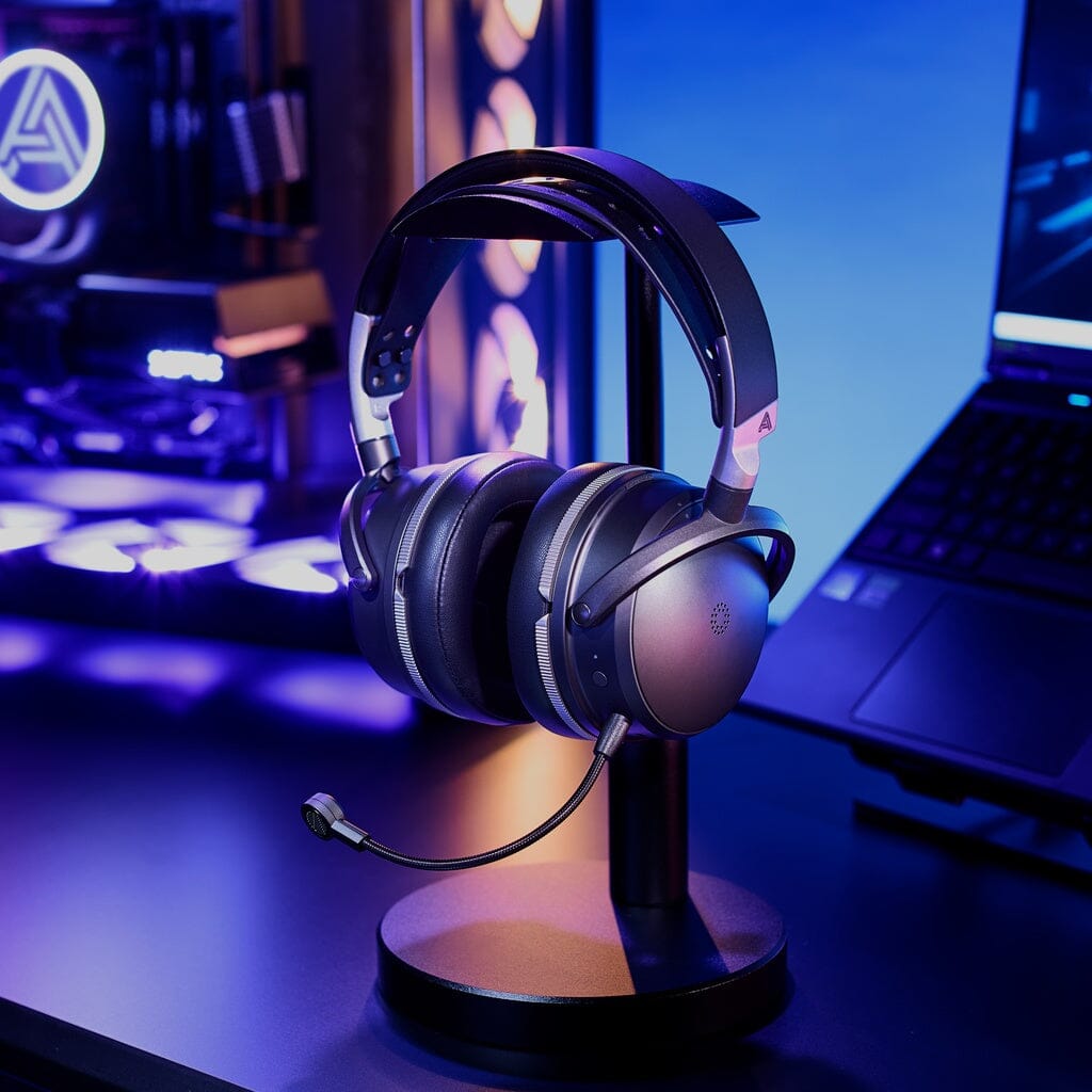 The Audeze Maxwell 2 ANC Wireless Headphones from Audeze, featuring a built-in microphone, rest on a stand placed on a dark desk next to a laptop and a desktop PC illuminated with vibrant LED lighting in a sleek modern setup.