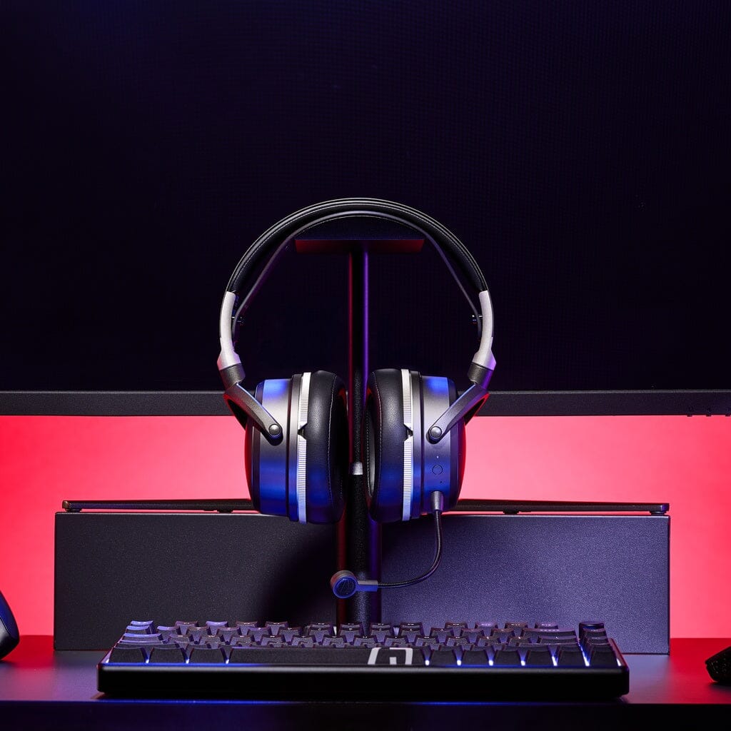 A pair of Audeze Maxwell 2 ANC Wireless Headphones by Audeze in black and silver rests on a stand behind a computer keyboard, set against a red backlit background.