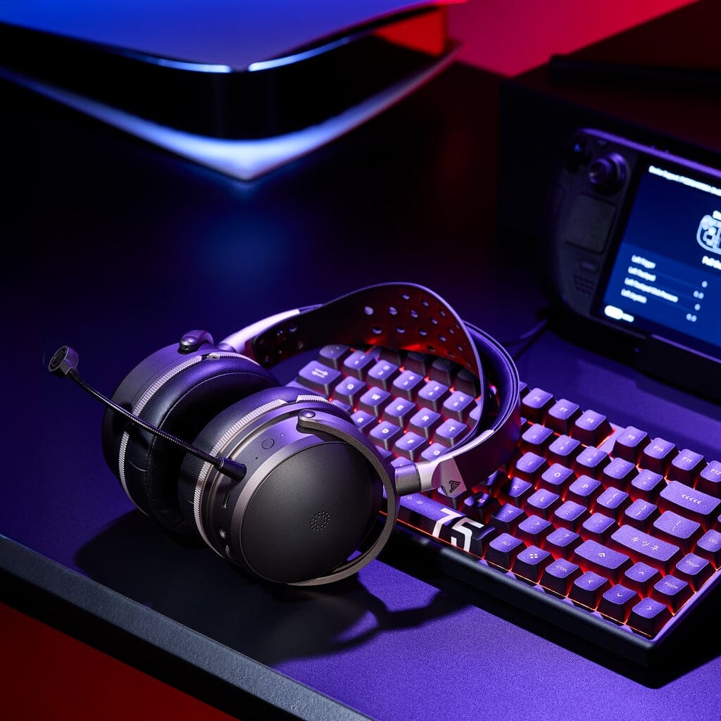 An Audeze Maxwell 2 ANC Wireless Headphones by Audeze, featuring a built-in microphone, rests on a mechanical keyboard. In the background, a handheld gaming console and part of another gaming console are visible, all bathed in purple and blue lighting.