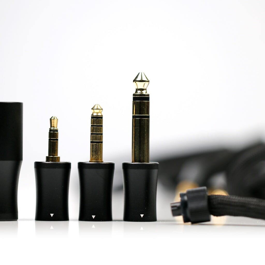 Three different sizes of gold-plated audio jack plugs with black bases are displayed upright in a row, with cables and connectors softly blurred in the background—ideal companions for your open-back headphone setup or flagship headphone models such as the Aune AR9000 Headphones from Aune Audio.