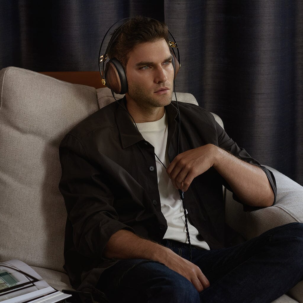 A man in a dark shirt sits on a beige armchair, focused, wearing Meze 99 Classics V2 headphones. One hand rests on his chest, the other holds the headphone cord. An open magazine lies beside him.