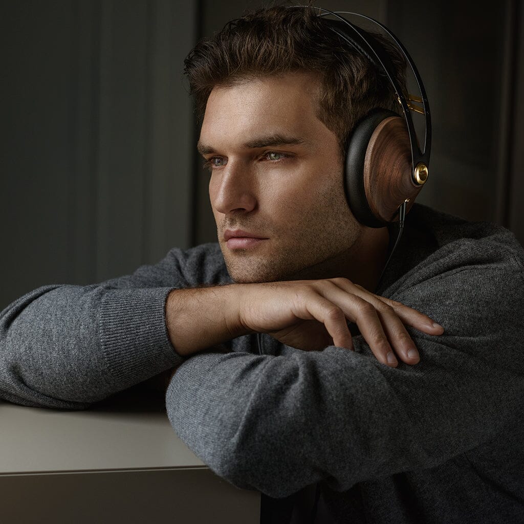 A man with short brown hair wearing Meze 99 Classics V2 headphones and a gray sweater rests his arms on a surface, gazing thoughtfully into the distance, enjoying their refined acoustic balance.