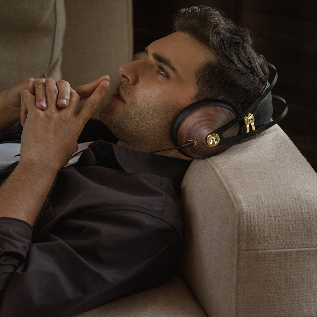 A man in a dark shirt relaxes thoughtfully on a beige couch, wearing Meze Audio 99 Classics V2 headphones by Meze, their walnut-wood and gold-accented design enhancing his enjoyment of the refined acoustic balance.
