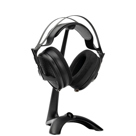 A pair of black over-ear headphones with a padded headband sits on the Meze Audio Manta Headphone Stand by Meze, featuring cable management, displayed against a white background.