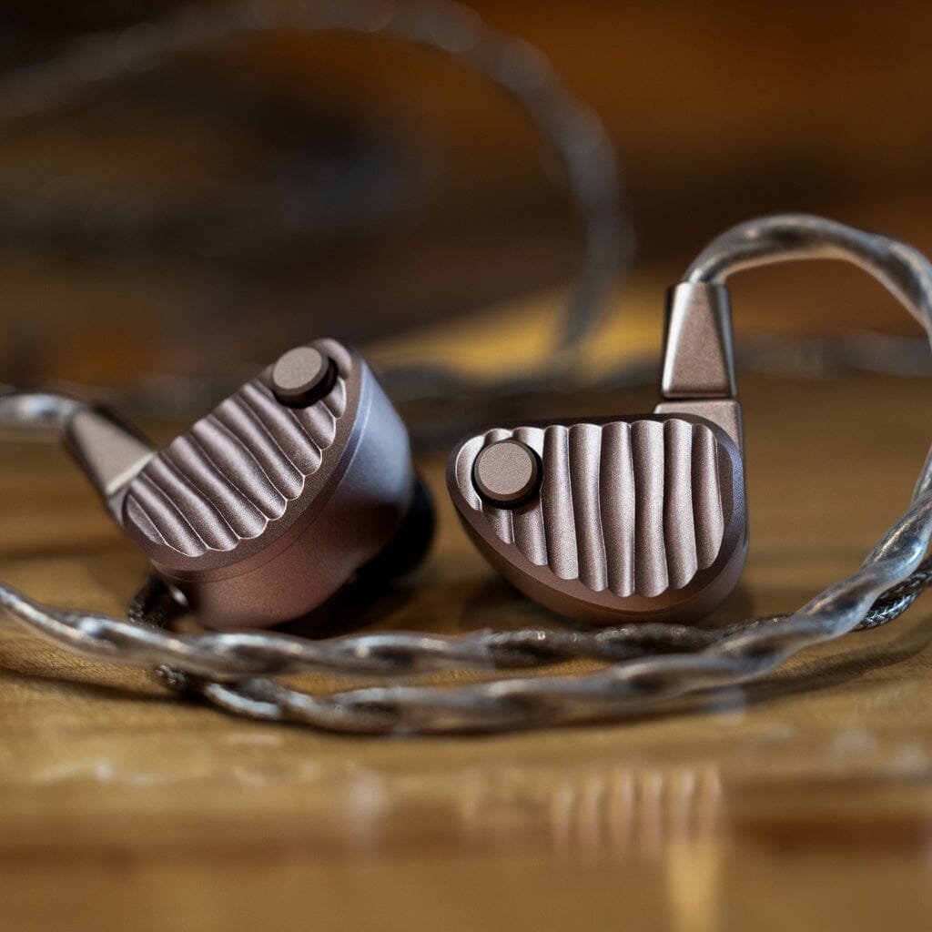 Two 64 Audio Ocean In-Ear Headphones with a ribbed metallic design rest on a wooden surface, partially encircled by their braided cable. The background is softly blurred in warm tones.