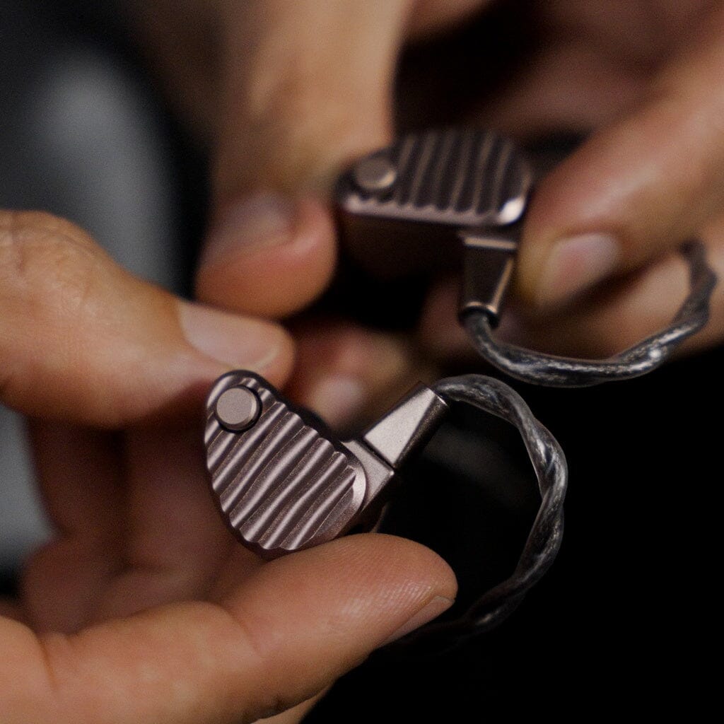 Close-up of hands holding a pair of 64 Audio Ocean In-Ear Headphones, the brand’s flagship metallic universal IEMs with a ridged texture and attached audio cables.