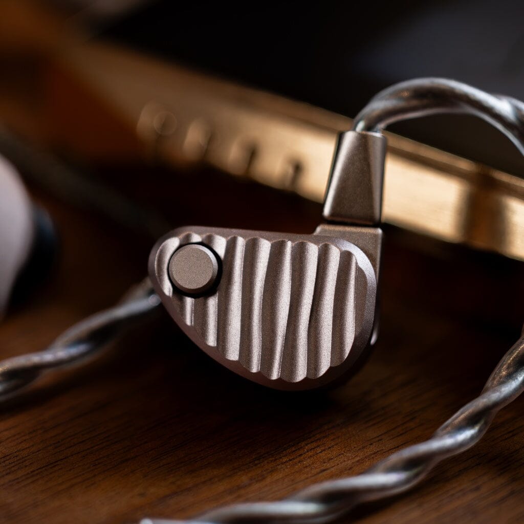 A close-up of the metallic, ridged 64 Audio Ocean In-Ear Headphones with a braided cable resting on a wooden surface, with a blurred electronic device in the background.