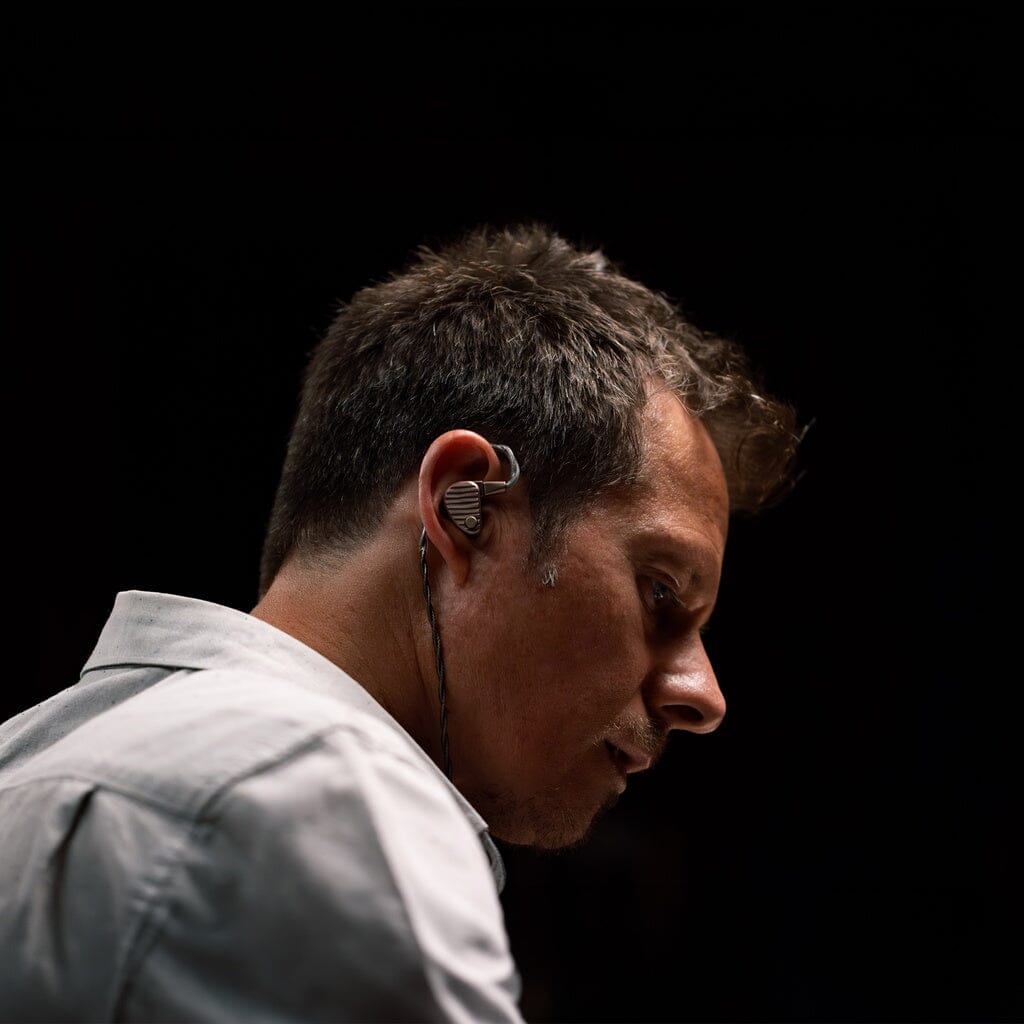 A man with short brown hair wears 64 Audio Ocean In-Ear Headphones by 64 Audio and looks down thoughtfully against a dark background.