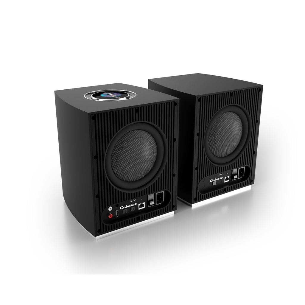 Two black Cabasse Rialto Wireless Powered Bookshelf Speakers are placed side by side on a white surface. The left speaker shows an exposed top section with internal components, while both feature front control panels and circular grilles.