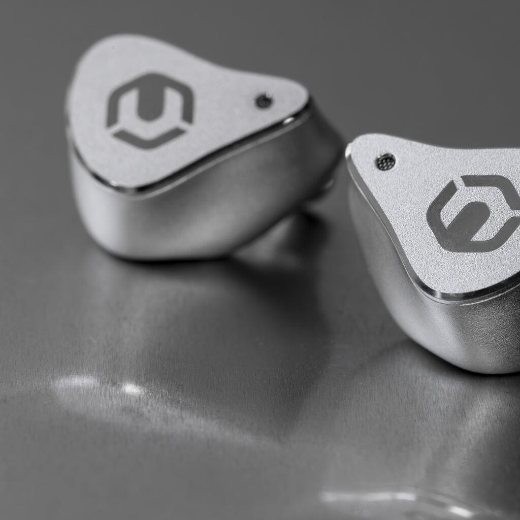 Close-up of two CrinEar Reference In-Ear Headphones by CrinEar, featuring a sleek silver finish and stylized logos, resting on a reflective gray surface—precision engineered for tonal accuracy.