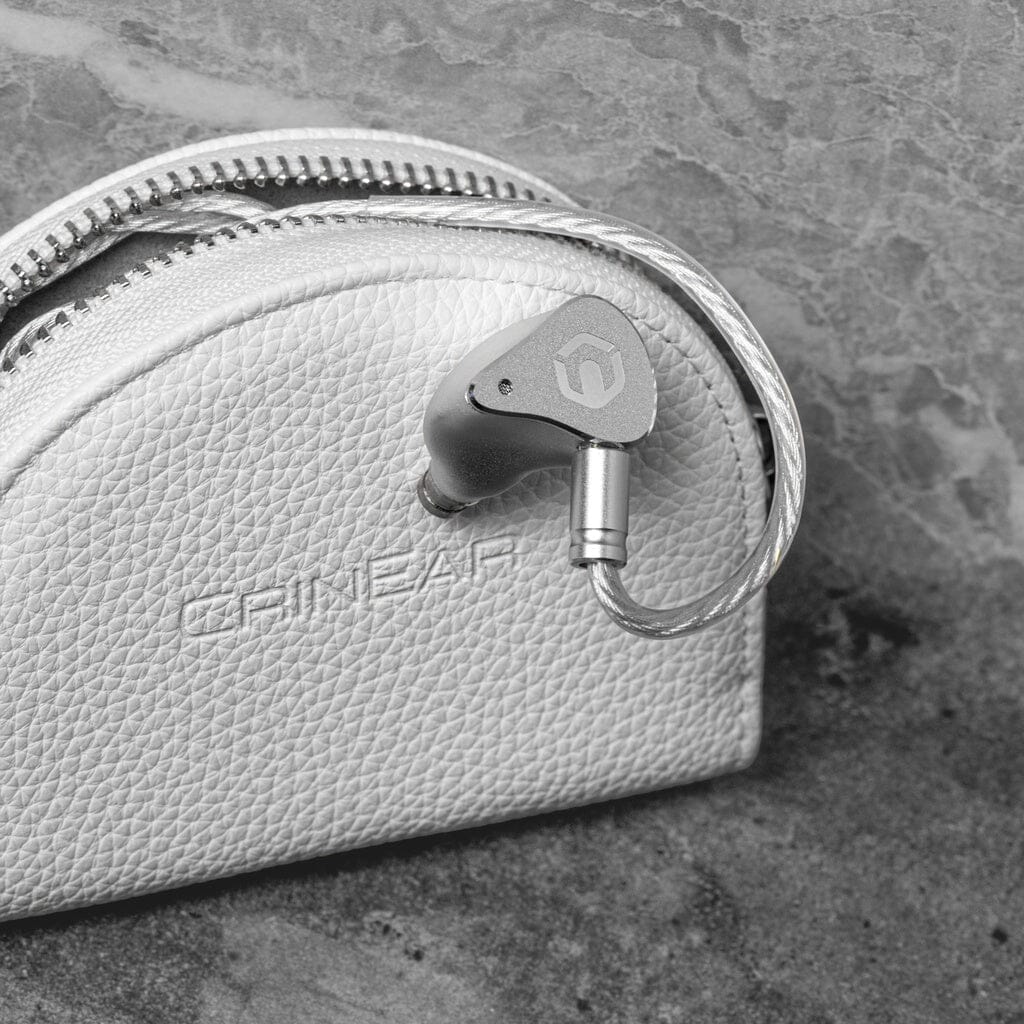 A metallic CrinEar Reference In-Ear Headphone with a detachable cable sits on a textured light leather zippered carrying case embossed with CRINEAR, set against a stone background, highlighting the headphone’s focus on tonal accuracy.