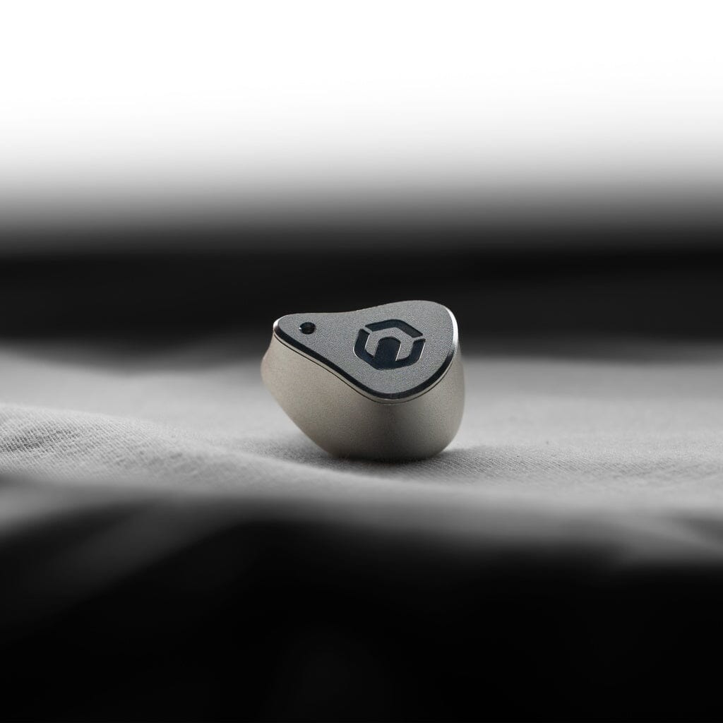 The CrinEar Reference In-Ear Headphones by CrinEar, featuring a metallic design and hexagon logo, rest on a soft white surface, offering tonal accuracy against a blurred black and white background.