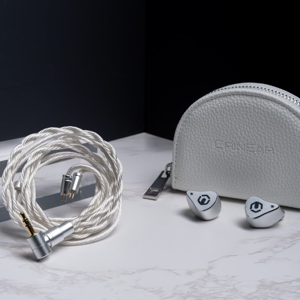A pair of CrinEar Reference In-Ear Headphones by CrinEar, a coiled white audio cable with a right-angle plug, and a white zippered carrying case are displayed on a white marble surface against a dark background.