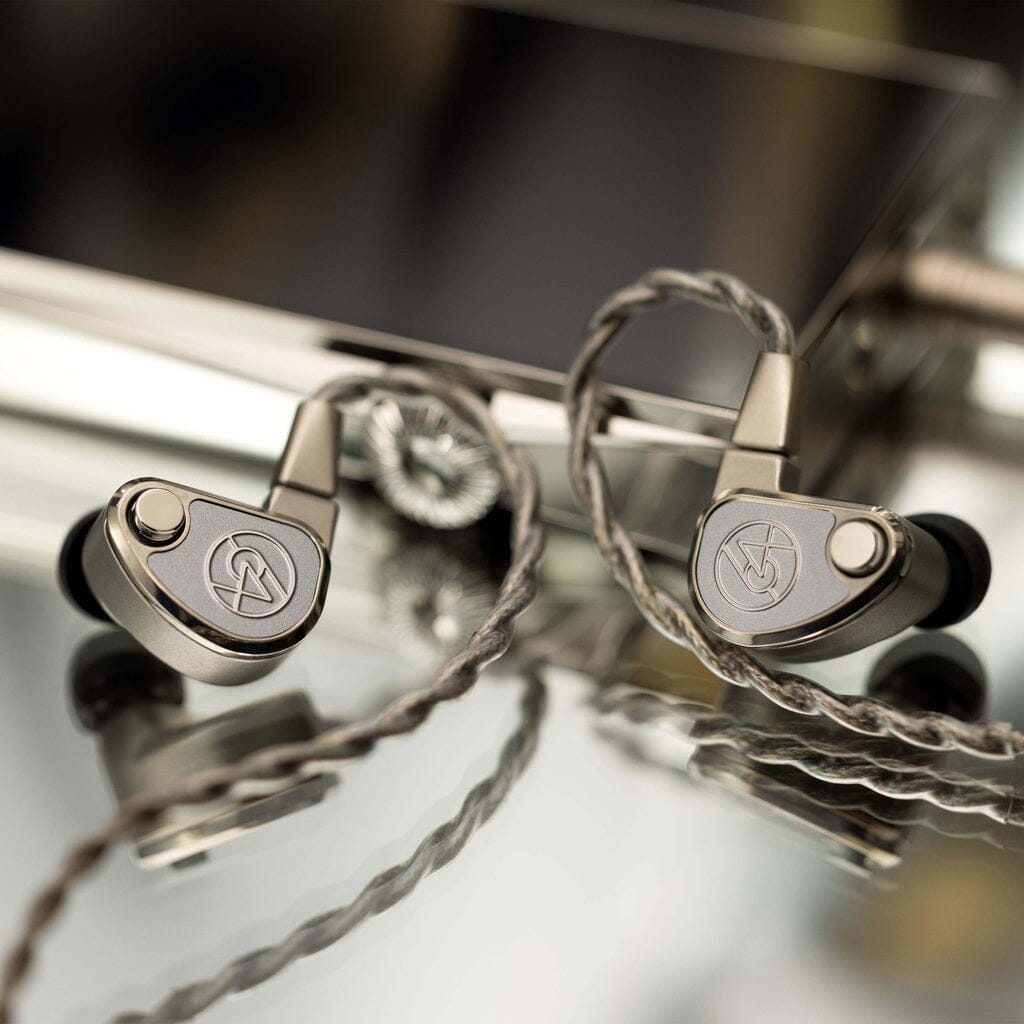 A pair of 64 Audio TWELVE Ti In-Ear Headphones by 64 Audio, featuring metallic earbuds with circular logos and braided cables, rest on a reflective surface with a blurred electronic device in the background.