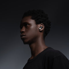 A young person with short curly hair wears a black T-shirt and Status Audio Pro X True Wireless - GoldenSound Edition earbuds, looking to the side against a dark gradient background. Preview
