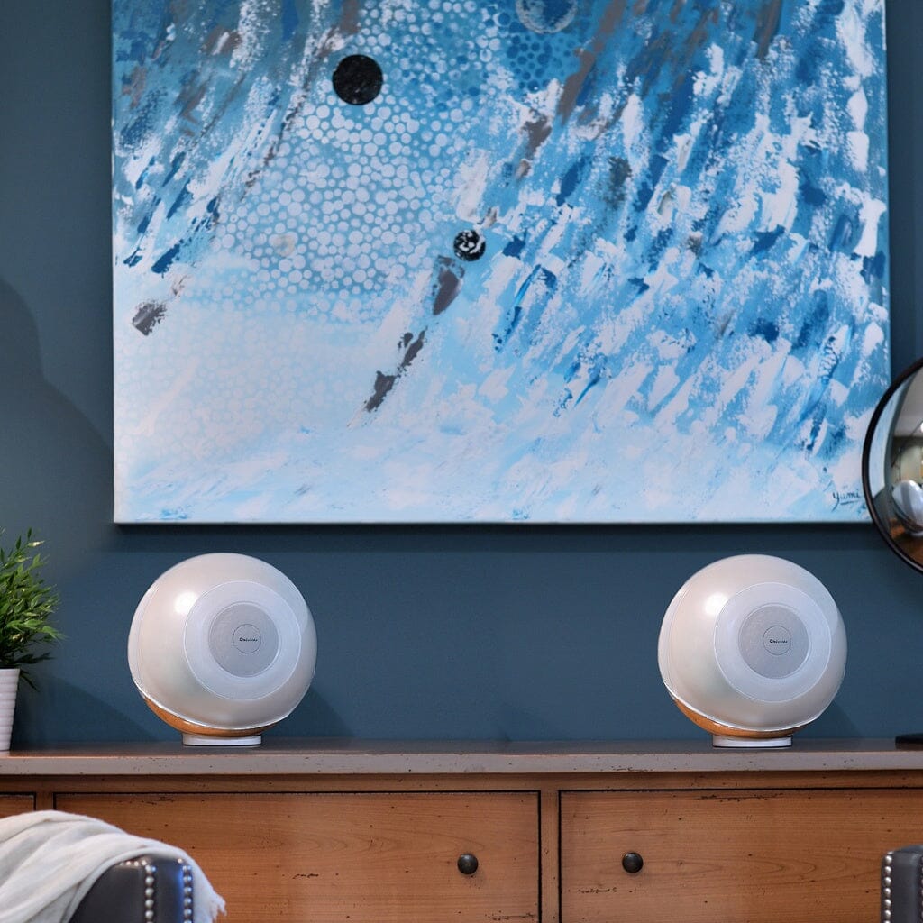 Two Cabasse THE PEARL AKOYA Wireless Speakers (pack of 2) sit on a wooden sideboard beneath an abstract blue and white painting in a modern room with blue walls, alongside a potted plant and part of a mirror.