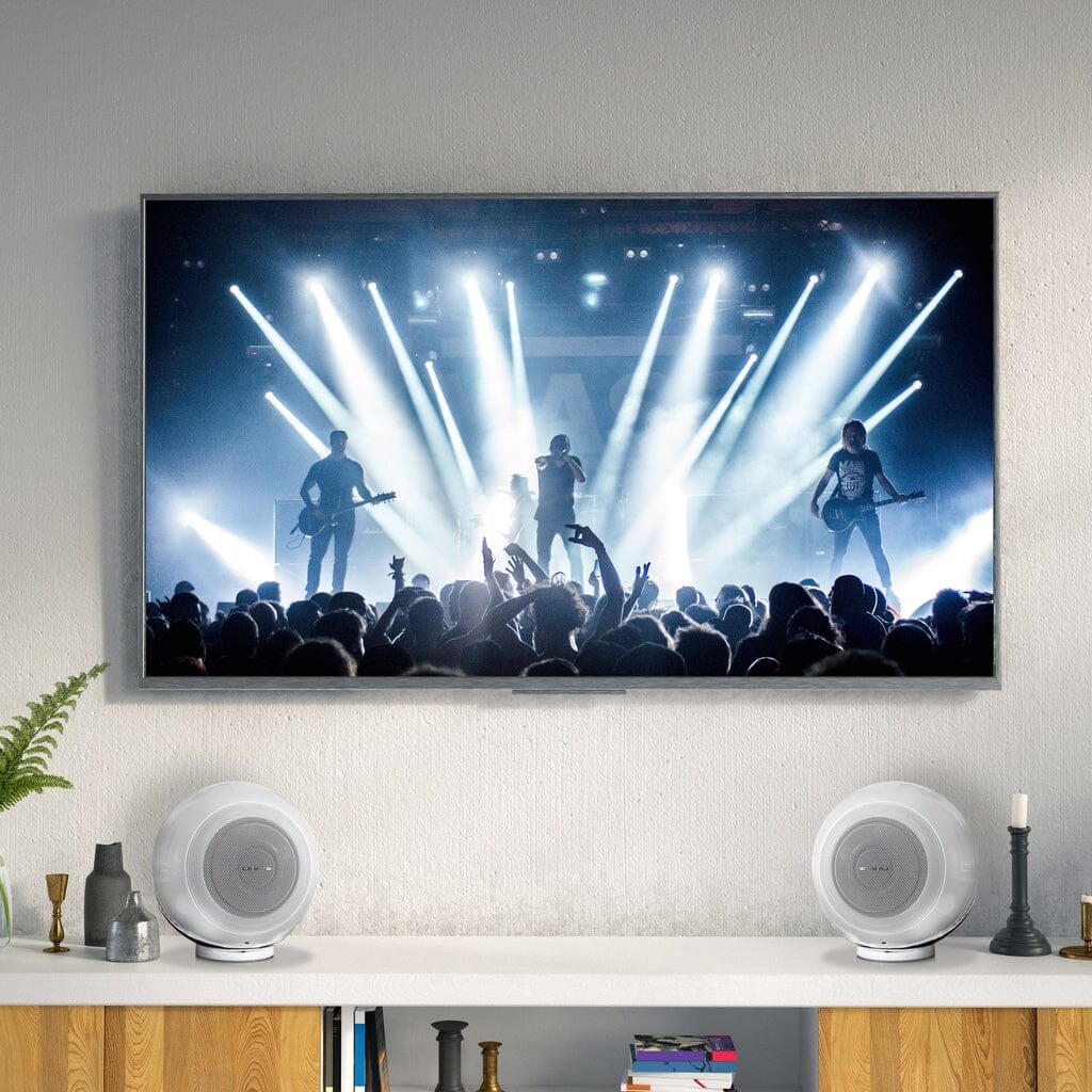 A wall-mounted TV shows a live concert, while Cabasse THE PEARL AKOYA Wireless Speaker - Pack of 2 delivers high-resolution audio from the shelf below, set among candles and decor.