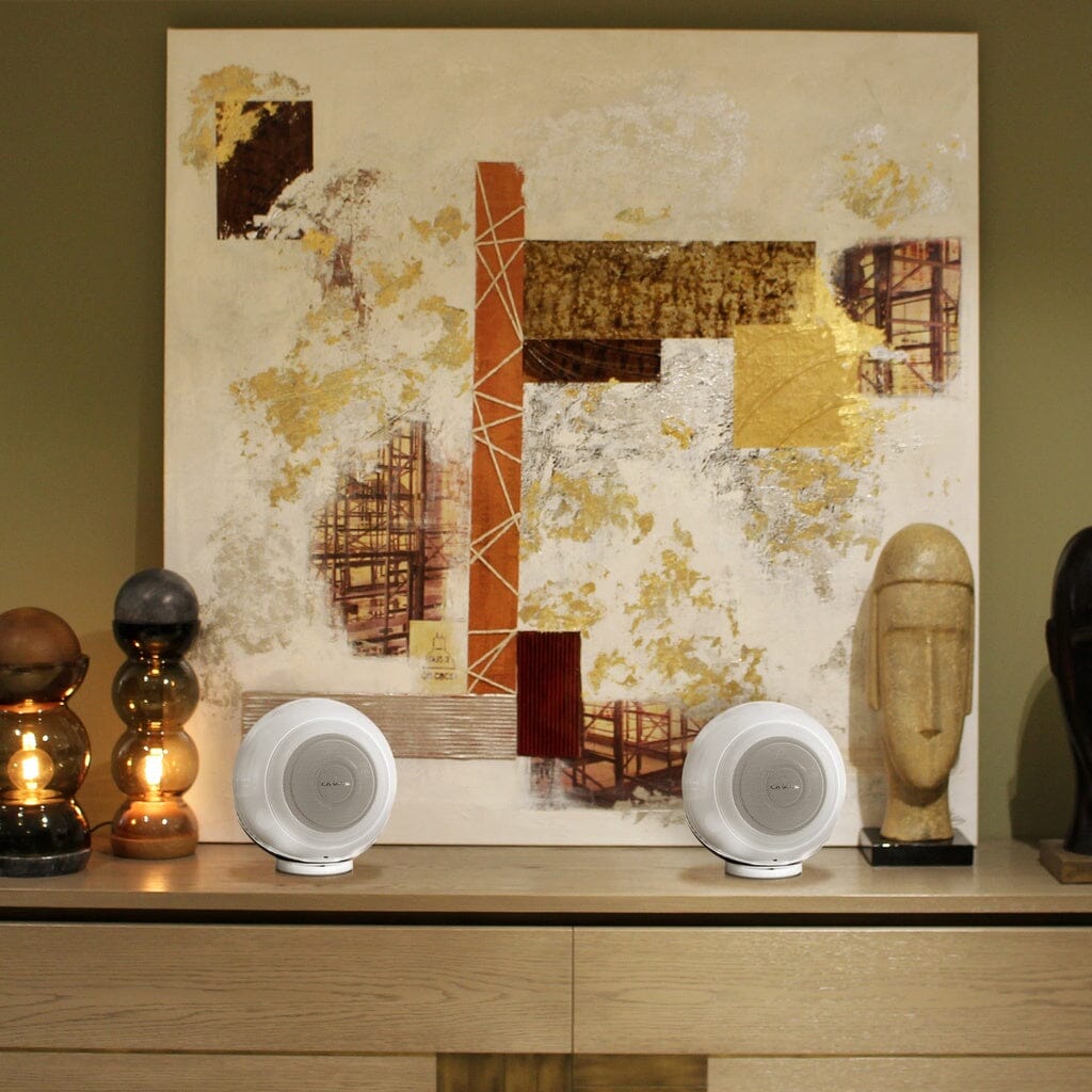 A wooden sideboard features a pair of Cabasse THE PEARL AKOYA wireless speakers, abstract art with orange and yellow accents, a stone bust, and two glass-based lamps as decorative highlights.