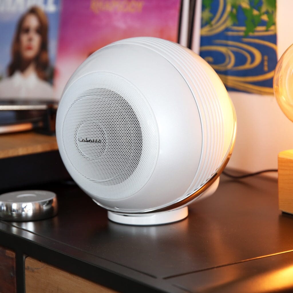 A pair of Cabasse THE PEARL AKOYA wireless speakers by Cabasse sit on a dark surface, with vinyl records and a lamp in the background, creating a modern and stylish audio setup.