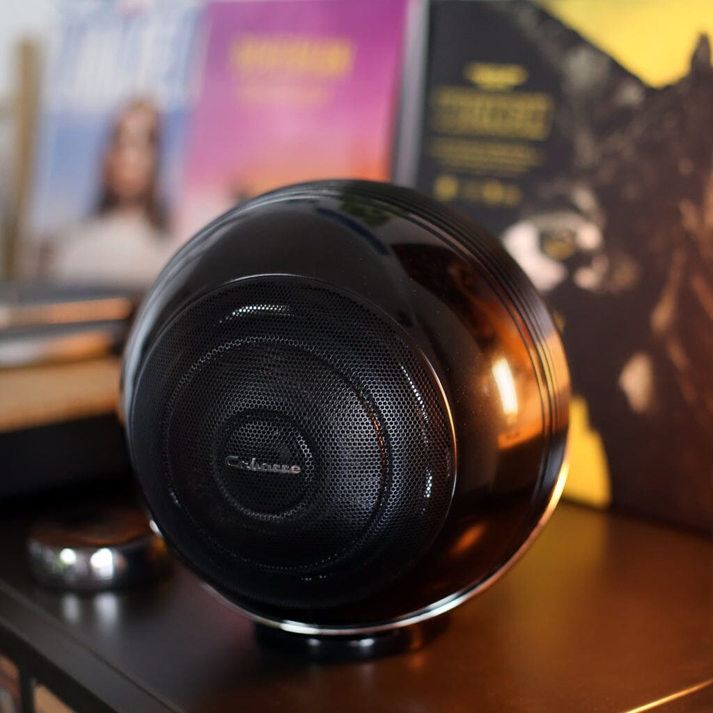 A close-up of Cabasse THE PEARL AKOYA Wireless Speaker – Pack of 2, featuring a tri-coaxial driver on a shelf, with blurred colorful album covers and books in the background. Warm lighting adds a cozy touch.