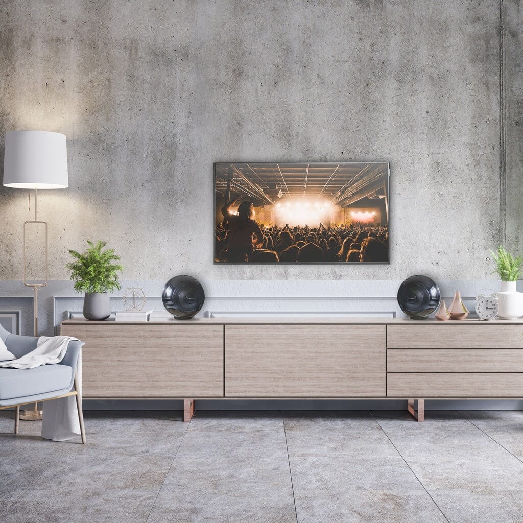 A modern living room with grey walls, light wood furniture, a white armchair, potted plants, and a pair of Cabasse THE PEARL AKOYA wireless speakers delivering hi-res audio beneath a large framed concert crowd photo above the sideboard.