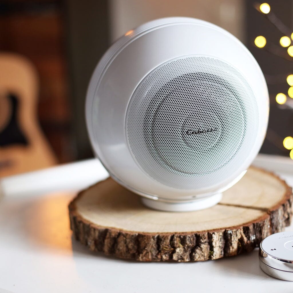 Two modern white Cabasse THE PEARL AKOYA wireless speakers sit on a round wooden slab on a table, with decorative lights and a guitar blurred in the background.