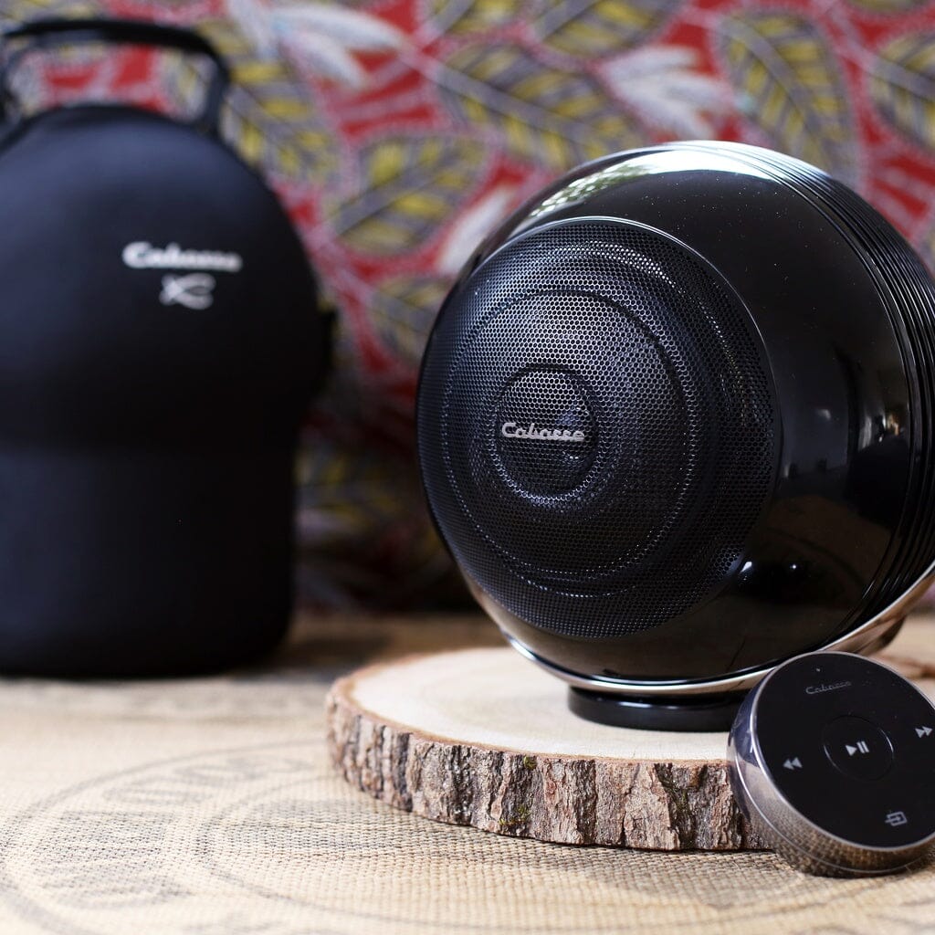 Two round, black Cabasse THE PEARL AKOYA wireless speakers sit on a wooden surface beside a small remote, with a carrying case and colorful patterned fabric in the background.
