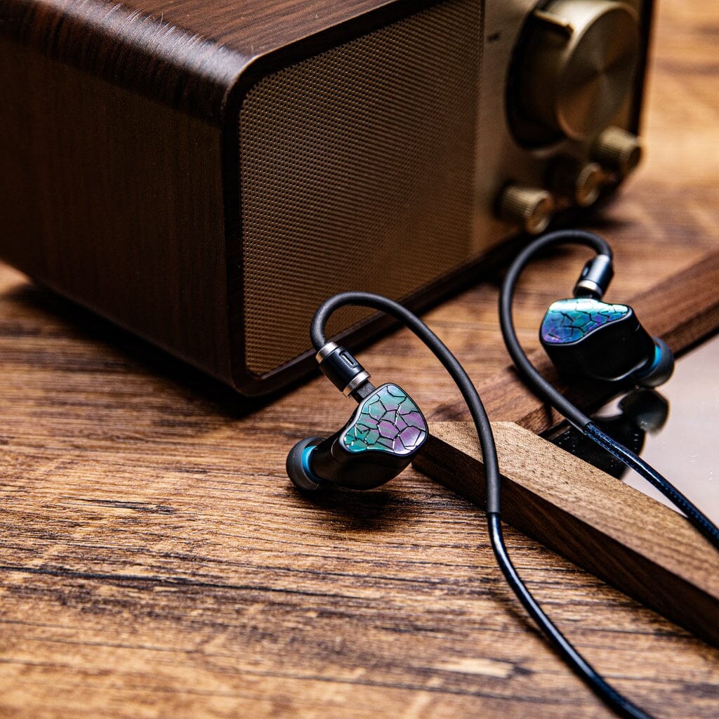 A pair of Thieaudio Hype 4 MK2 in-ear headphones with colorful, cracked-pattern faceplates rest on a wooden surface beside a vintage-style wooden radio and the corner of a smartphone.