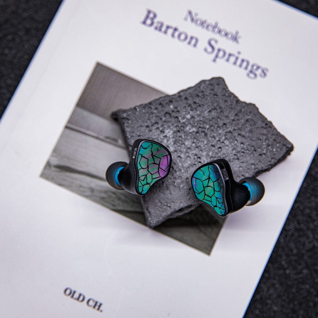 A pair of Thieaudio Hype 4 MK2 In-Ear Headphones with a green and purple mosaic design rests on textured gray stone, set atop the book "Notebook Barton Springs" by OLD CH.