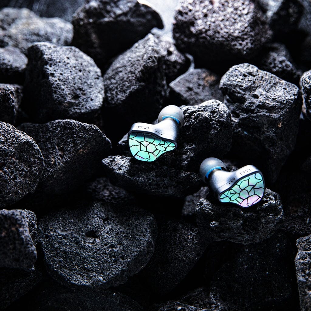 A pair of Thieaudio Hype 4 MK2 In-Ear Headphones with iridescent, cracked glass-style surfaces rest on rough, dark volcanic rocks.
