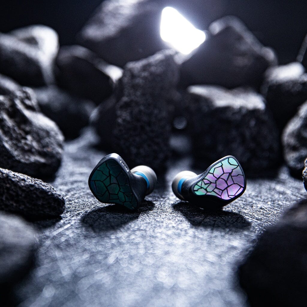 The Thieaudio Hype 4 MK2 In-Ear Headphones, by Thieaudio, showcase green and pink mosaic-patterned designs resting on a textured surface among dark rocks, with dramatic backlighting emphasizing their bold look.