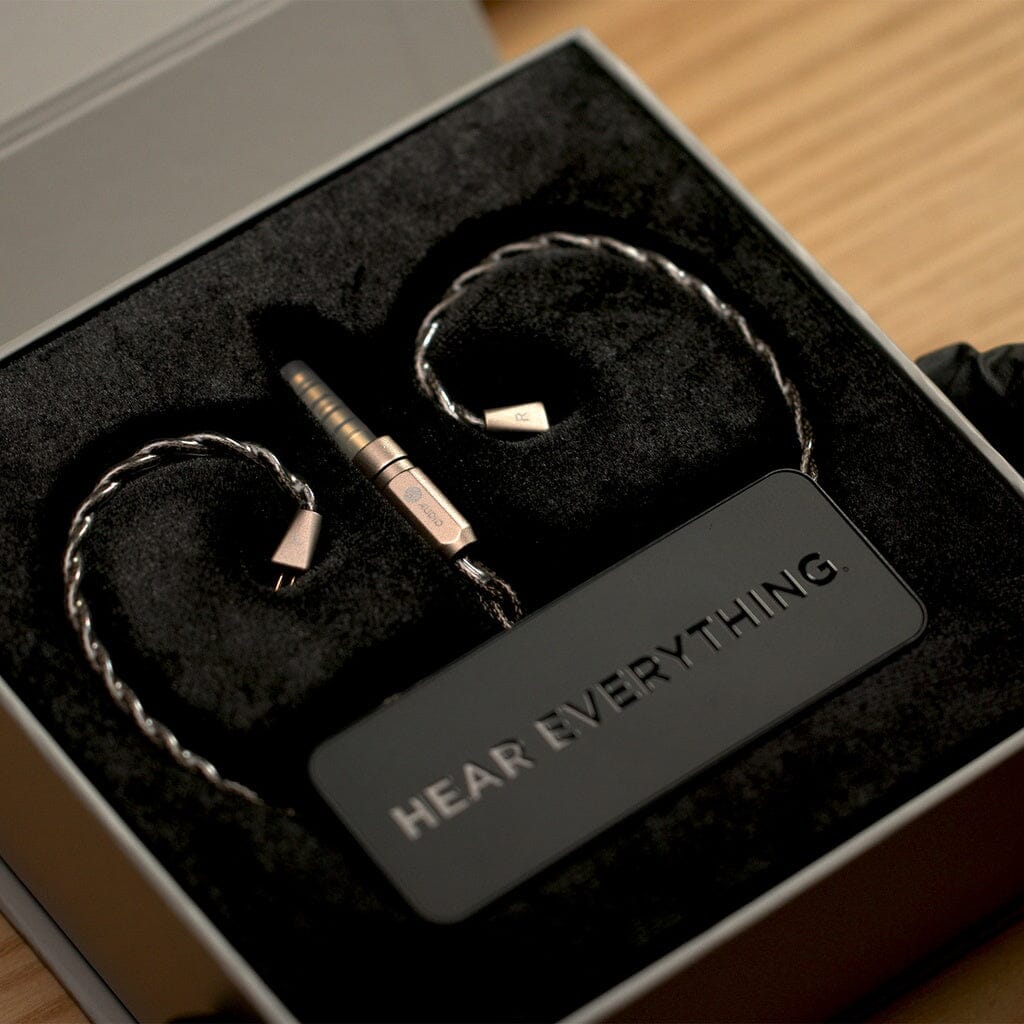 A pair of 64 Audio TWELVE Ti In-Ear Headphones with braided cables sit in a black foam case next to a tag reading "HEAR EVERYTHING," all from 64 Audio, displayed inside an open box on a wooden surface.